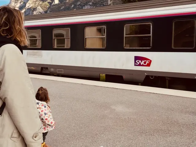 Une femme et un enfant attendent un train de nuit devant un train SNCF avec des montagnes en arrière-plan