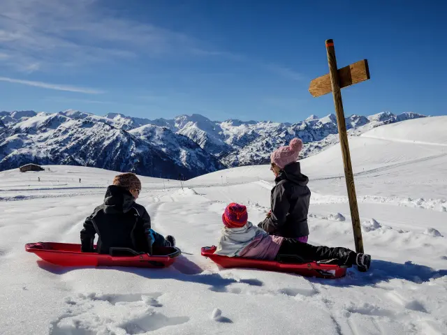 Trois enfants en luge sur une pente enneigée avec des montagnes en arrière-plan