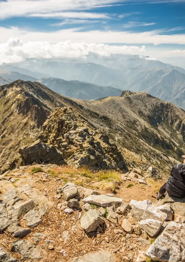 Deux randonneurs assis au sommet du Canigó contemplent la chaîne montagneuse des Pyrénées sous un ciel dégagé.