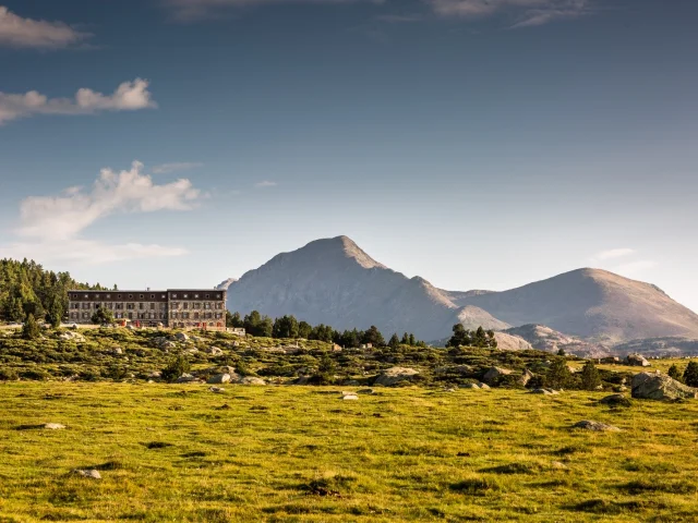 Bâtiment de montagne au pied du massif du Carlit dans les Pyrénées-Orientales, entouré de forêts et de prairies d’altitude sous un ciel clair.