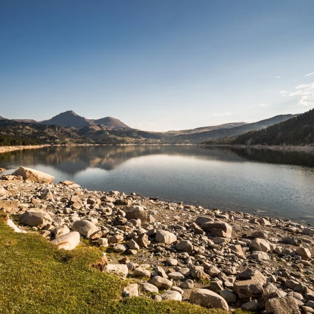 Lac de montagne entouré de forêts et de sommets dans les Pyrénées-Orientales, sous un ciel bleu dégagé.