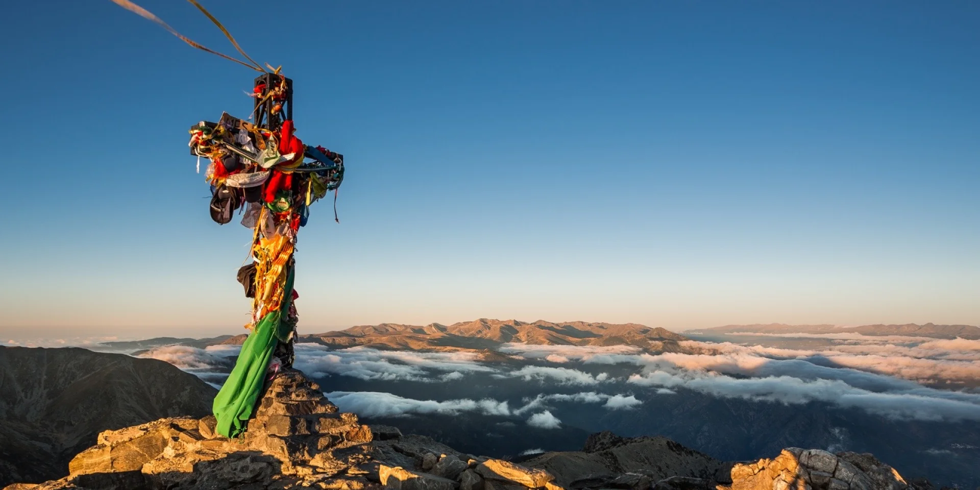 Croix du sommet du Canigó ornée de rubans colorés, dominant les montagnes et les nuages au lever du soleil dans les Pyrénées-Orientales.
