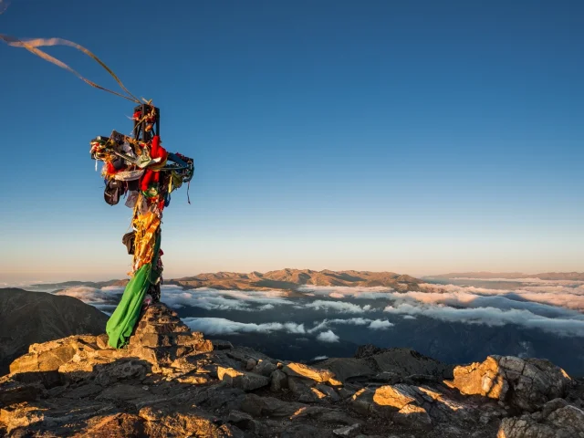Croix du sommet du Canigó ornée de rubans colorés, dominant les montagnes et les nuages au lever du soleil dans les Pyrénées-Orientales.