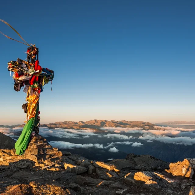 Croix du sommet du Canigó ornée de rubans colorés, dominant les montagnes et les nuages au lever du soleil dans les Pyrénées-Orientales.