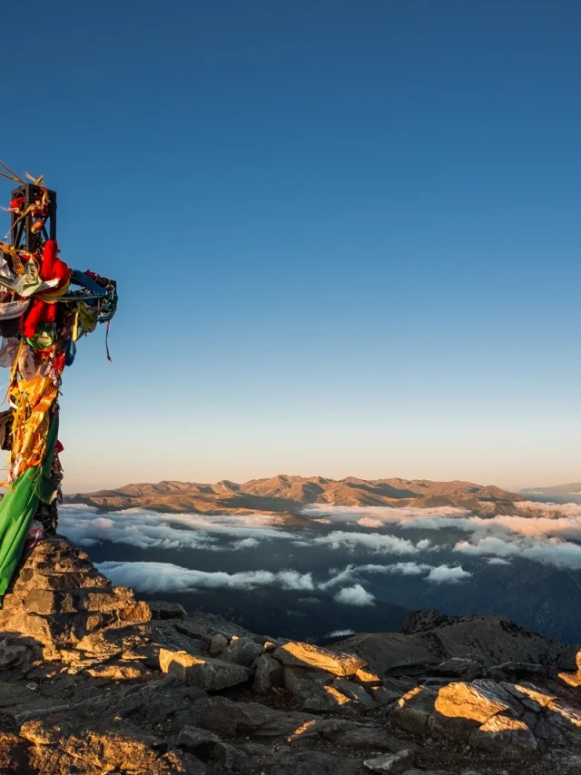 Croix du sommet du Canigó ornée de rubans colorés, dominant les montagnes et les nuages au lever du soleil dans les Pyrénées-Orientales.