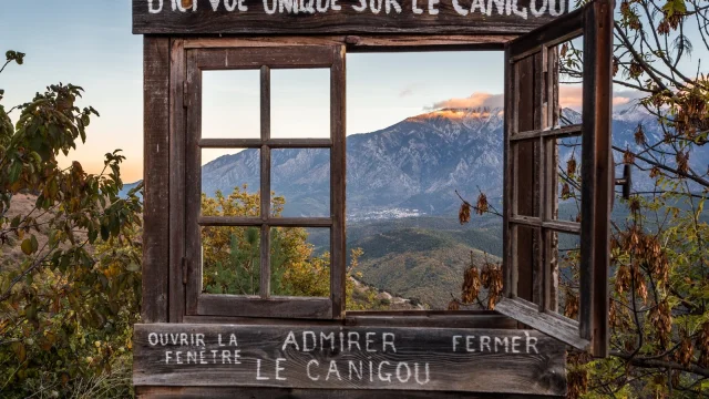 Fenêtre en bois encadrant une vue sur le massif du Canigó dans les Pyrénées-Orientales, avec l’inscription “D’ici vue unique sur le Canigou”.