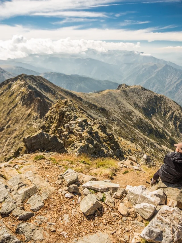 Deux randonneurs assis au sommet du Canigó contemplent la chaîne montagneuse des Pyrénées sous un ciel dégagé.