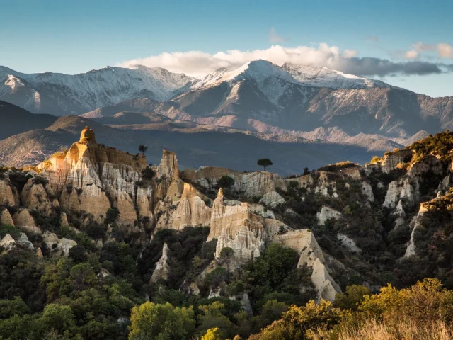 Les orgues d’Ille-sur-Têt dans les Pyrénées-Orientales, formations d’argile et de sable au premier plan, avec le massif du Canigó enneigé en arrière-plan sous un ciel bleu.