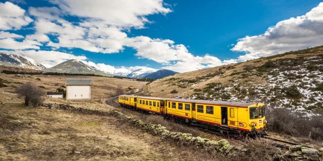 Le Train Jaune traverse un paysage de montagne, entre prairies et sommets enneigés, sous un ciel bleu parsemé de nuages.