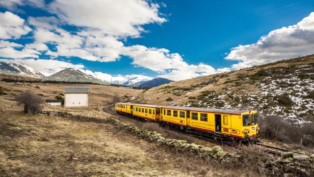 Le Train Jaune traverse un paysage de montagne, entre prairies et sommets enneigés, sous un ciel bleu parsemé de nuages.