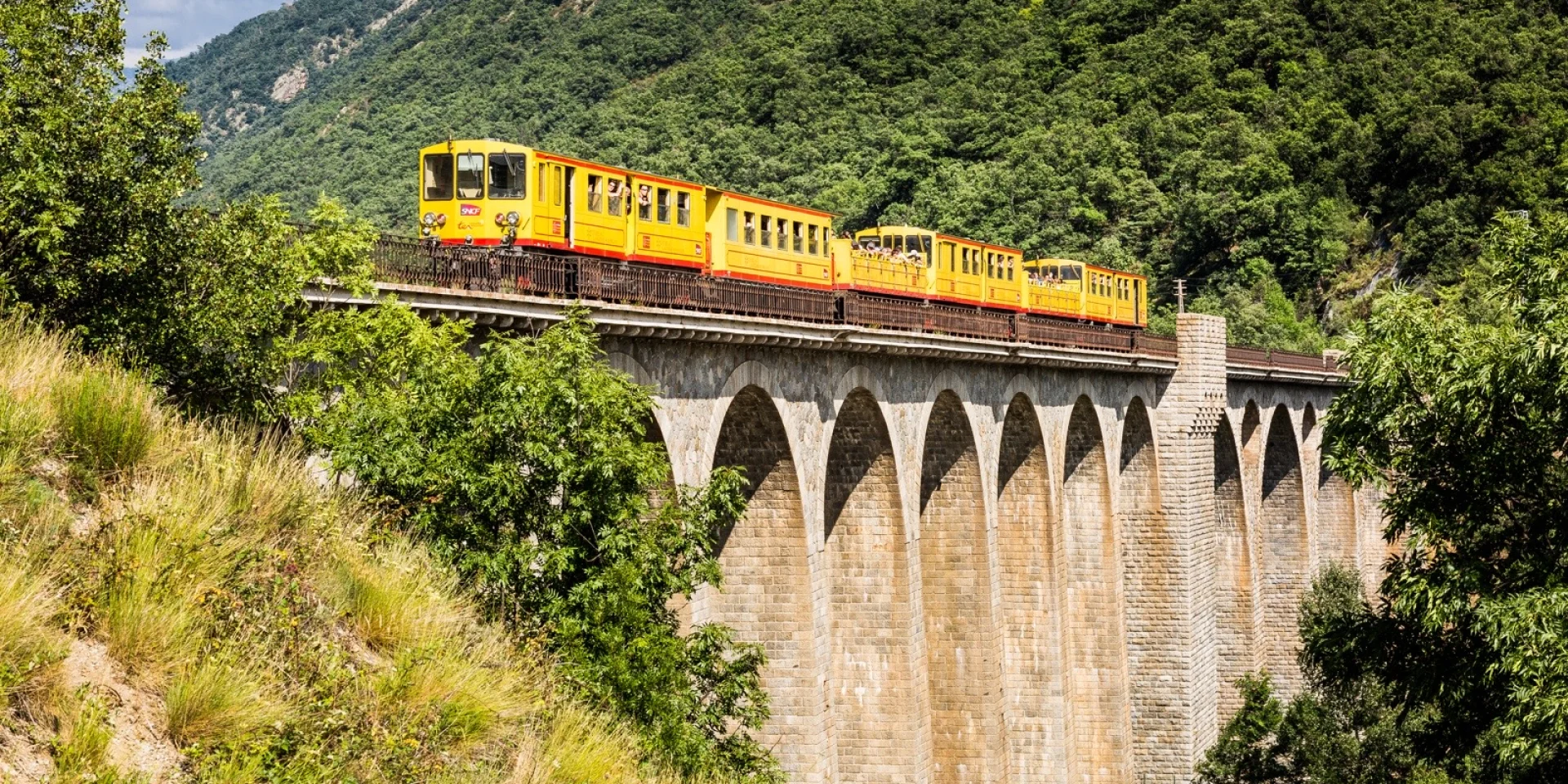 Le Train Jaune traverse un grand viaduc en pierre au cœur d’un paysage de montagnes verdoyantes dans les Pyrénées-Orientales.