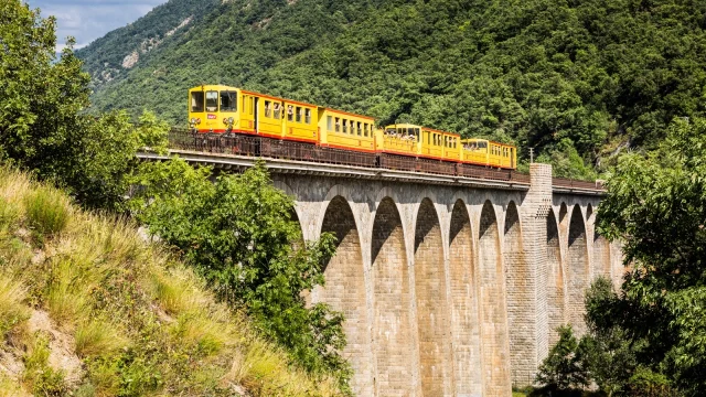 Le Train Jaune traverse un grand viaduc en pierre au cœur d’un paysage de montagnes verdoyantes dans les Pyrénées-Orientales.