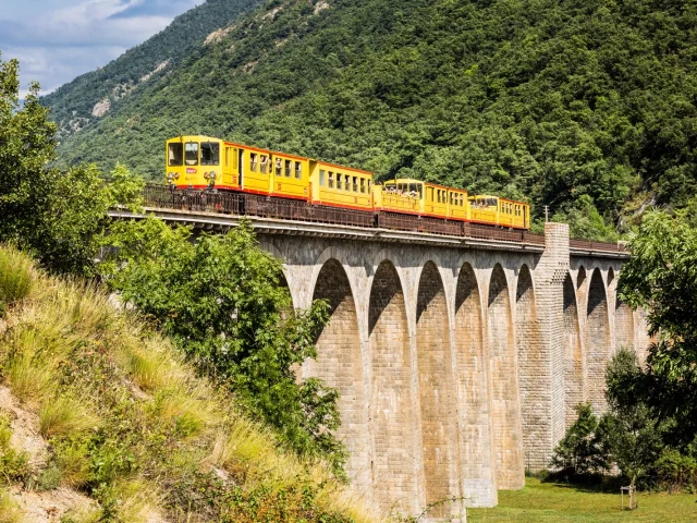 Le Train Jaune traverse un grand viaduc en pierre au cœur d’un paysage de montagnes verdoyantes dans les Pyrénées-Orientales.