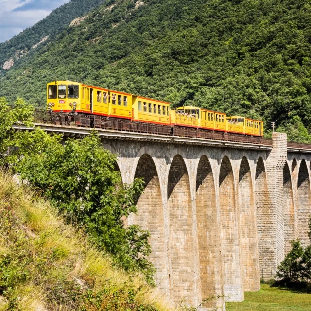Le Train Jaune traverse un grand viaduc en pierre au cœur d’un paysage de montagnes verdoyantes dans les Pyrénées-Orientales.