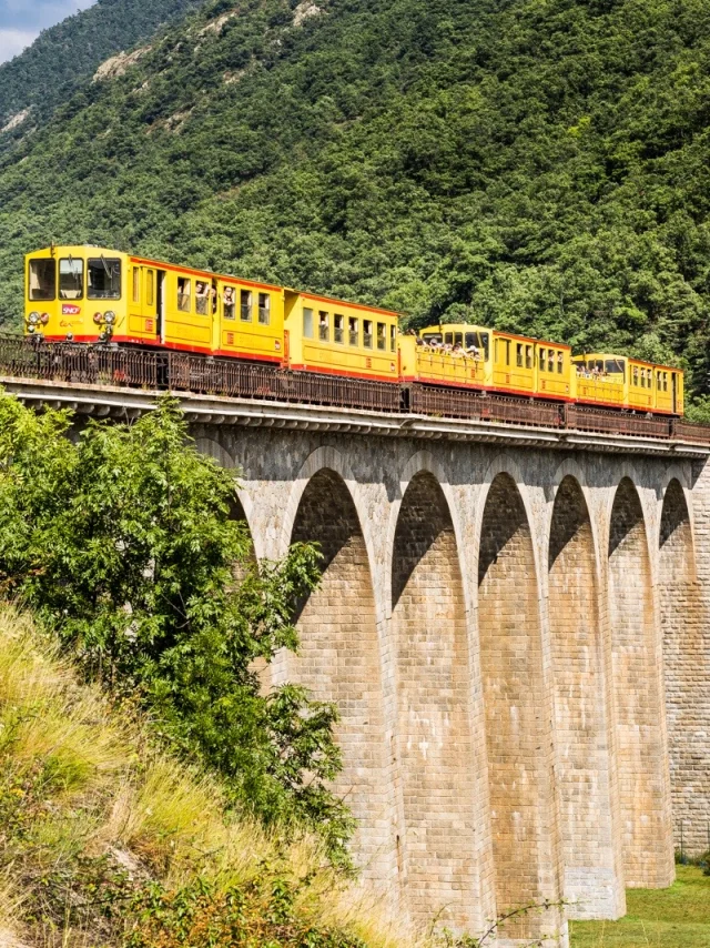 Le Train Jaune traverse un grand viaduc en pierre au cœur d’un paysage de montagnes verdoyantes dans les Pyrénées-Orientales.