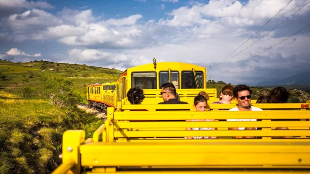 Voyageurs à bord du Train Jaune dans un wagon découvert, traversant les paysages verdoyants des Pyrénées-Orientales sous un ciel partiellement nuageux.