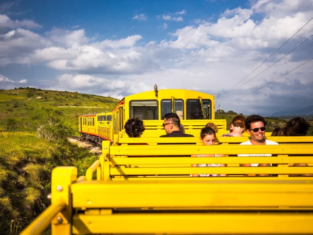 Voyageurs à bord du Train Jaune dans un wagon découvert, traversant les paysages verdoyants des Pyrénées-Orientales sous un ciel partiellement nuageux.