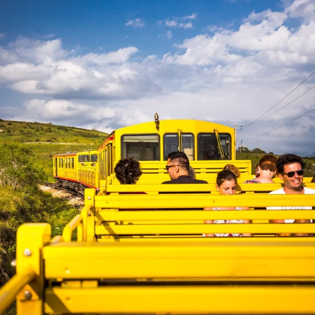 Voyageurs à bord du Train Jaune dans un wagon découvert, traversant les paysages verdoyants des Pyrénées-Orientales sous un ciel partiellement nuageux.