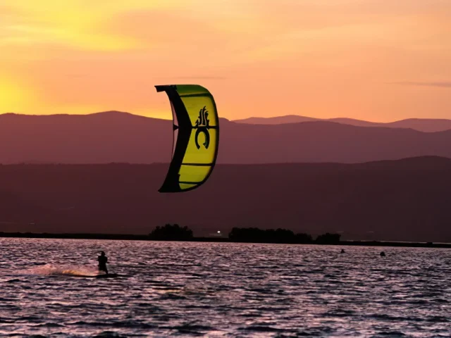 Personne pratiquant le kitesurf au coucher du soleil sur la mer, avec les montagnes en arrière-plan.