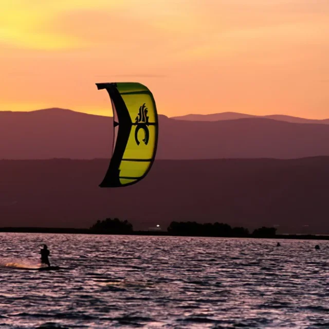 Personne pratiquant le kitesurf au coucher du soleil sur la mer, avec les montagnes en arrière-plan.