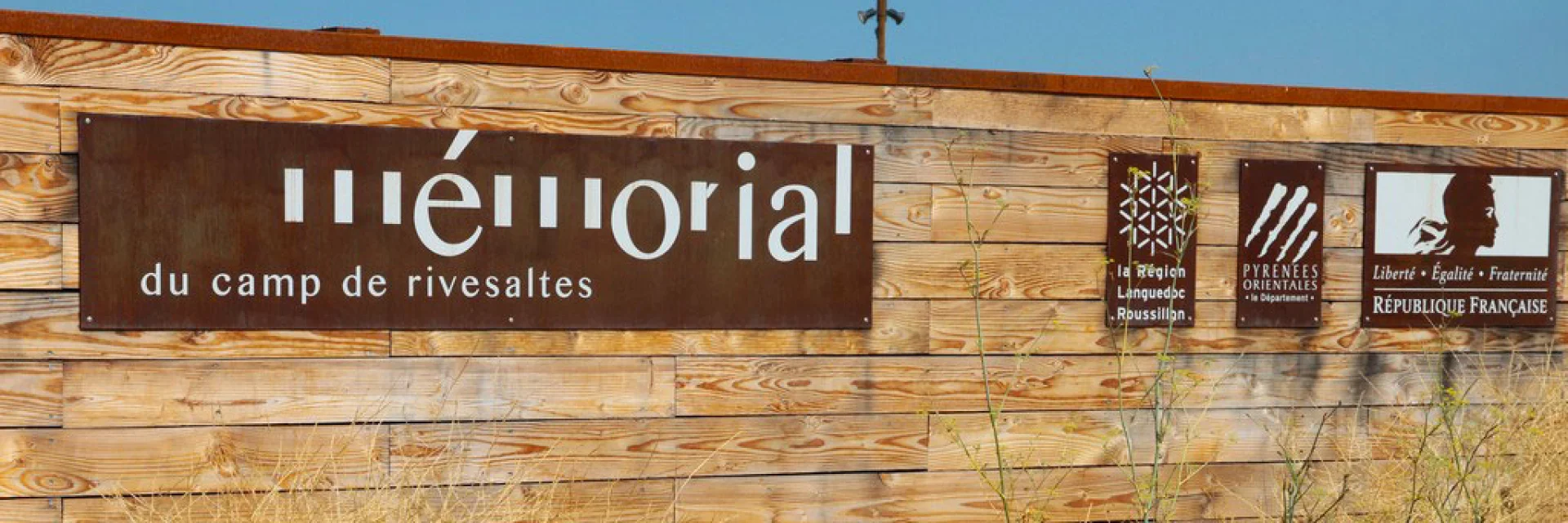 Panneau d’entrée du Mémorial du Camp de Rivesaltes, fixé sur une palissade en bois, avec les logos de la Région Languedoc-Roussillon, du Département des Pyrénées-Orientales et de la République Française.
