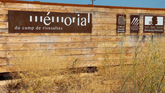Panneau d’entrée du Mémorial du Camp de Rivesaltes, fixé sur une palissade en bois, avec les logos de la Région Languedoc-Roussillon, du Département des Pyrénées-Orientales et de la République Française.