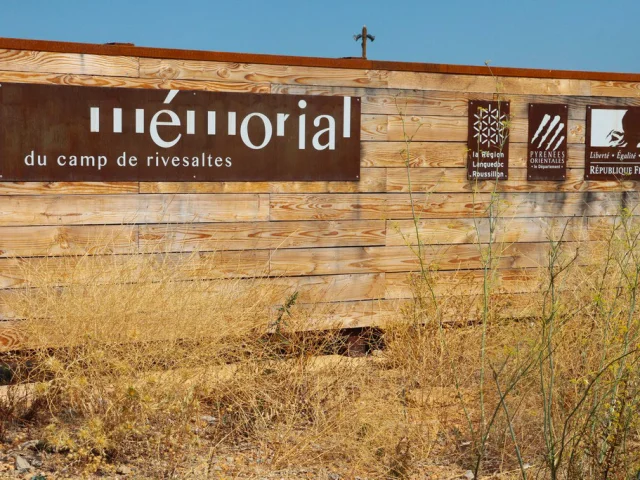 Panneau d’entrée du Mémorial du Camp de Rivesaltes, fixé sur une palissade en bois, avec les logos de la Région Languedoc-Roussillon, du Département des Pyrénées-Orientales et de la République Française.