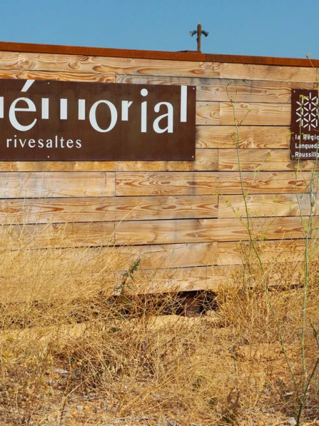 Panneau d’entrée du Mémorial du Camp de Rivesaltes, fixé sur une palissade en bois, avec les logos de la Région Languedoc-Roussillon, du Département des Pyrénées-Orientales et de la République Française.