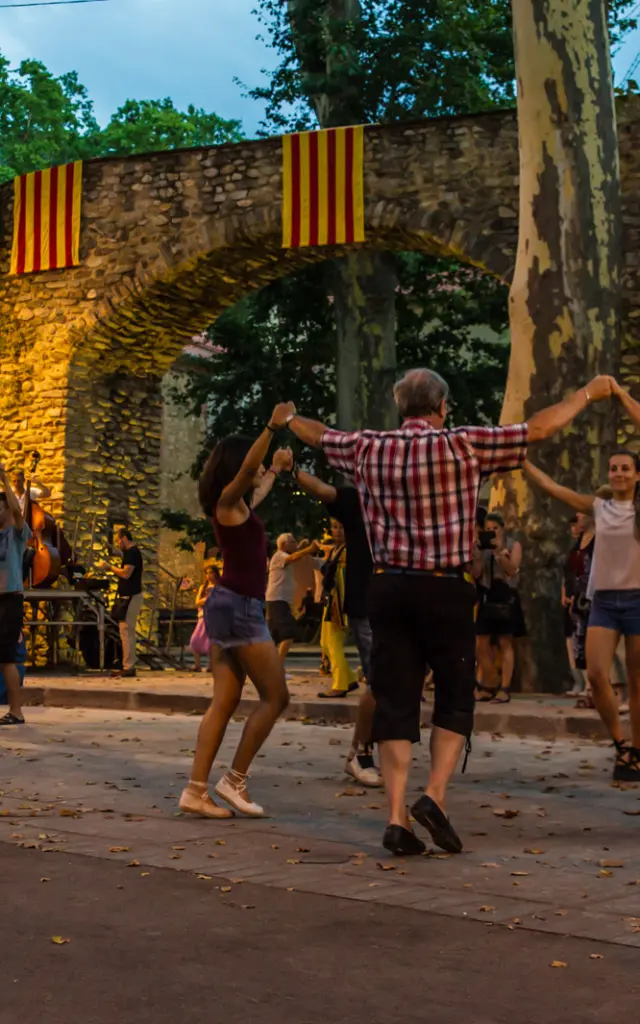 Groupe de personnes dansant la sardane en cercle sur une place de Céret, devant un orchestre de cobla et des arcades en pierre décorées de drapeaux catalans.