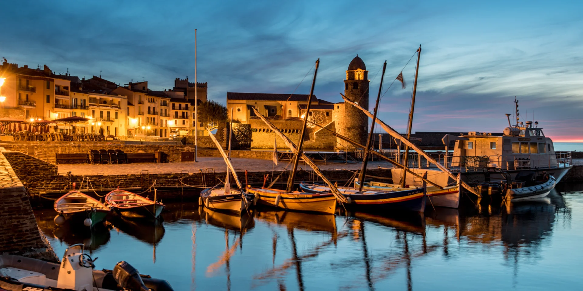 Vue du port de Collioure à la tombée de la nuit, avec les barques catalanes amarrées et l’église Notre-Dame-des-Anges illuminée en arrière-plan.