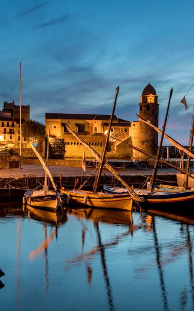 Vue du port de Collioure à la tombée de la nuit, avec les barques catalanes amarrées et l’église Notre-Dame-des-Anges illuminée en arrière-plan.