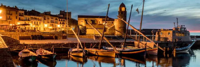 Vue du port de Collioure à la tombée de la nuit, avec les barques catalanes amarrées et l’église Notre-Dame-des-Anges illuminée en arrière-plan.