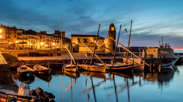 Vue du port de Collioure à la tombée de la nuit, avec les barques catalanes amarrées et l’église Notre-Dame-des-Anges illuminée en arrière-plan.