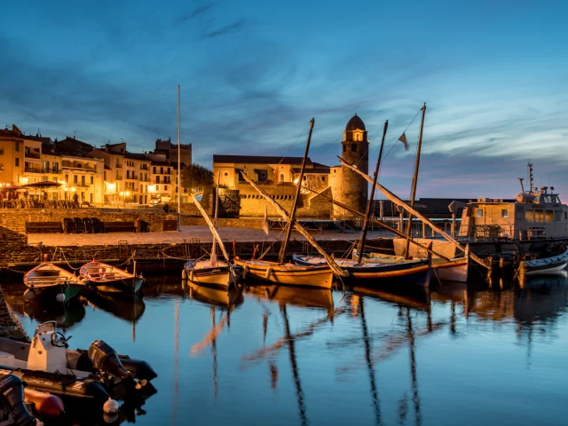 Vue du port de Collioure à la tombée de la nuit, avec les barques catalanes amarrées et l’église Notre-Dame-des-Anges illuminée en arrière-plan.
