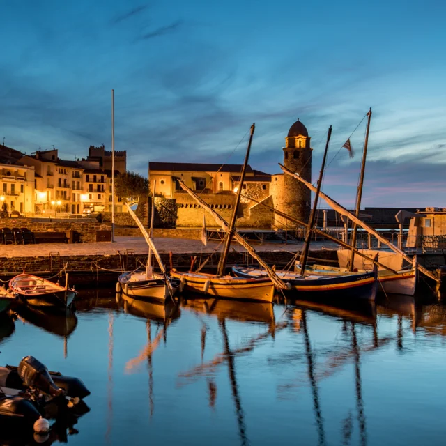Vue du port de Collioure à la tombée de la nuit, avec les barques catalanes amarrées et l’église Notre-Dame-des-Anges illuminée en arrière-plan.