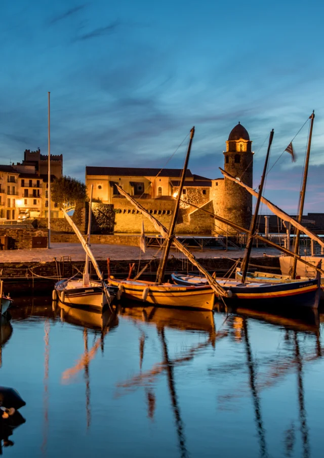 Vue du port de Collioure à la tombée de la nuit, avec les barques catalanes amarrées et l’église Notre-Dame-des-Anges illuminée en arrière-plan.