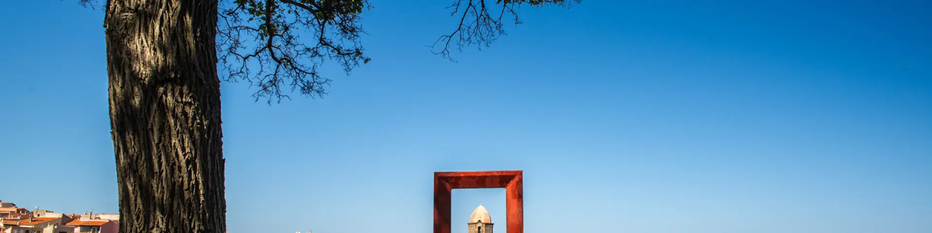 Vue sur le clocher de l’église Notre-Dame-des-Anges de Collioure encadré par une sculpture carrée en métal rouge, avec la mer, les voiliers et les maisons colorées du port en arrière-plan.