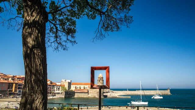 Vue sur le clocher de l’église Notre-Dame-des-Anges de Collioure encadré par une sculpture carrée en métal rouge, avec la mer, les voiliers et les maisons colorées du port en arrière-plan.
