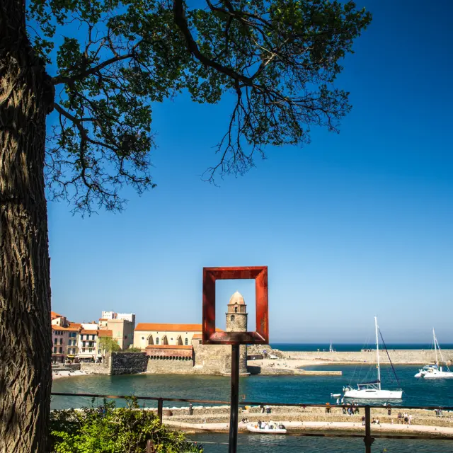 Vue sur le clocher de l’église Notre-Dame-des-Anges de Collioure encadré par une sculpture carrée en métal rouge, avec la mer, les voiliers et les maisons colorées du port en arrière-plan.
