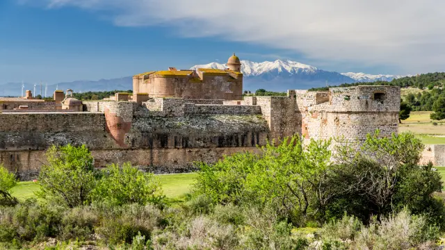 Vue de la forteresse de Salses, imposante construction en briques et pierres, avec le Canigó enneigé en arrière-plan sous un ciel bleu.