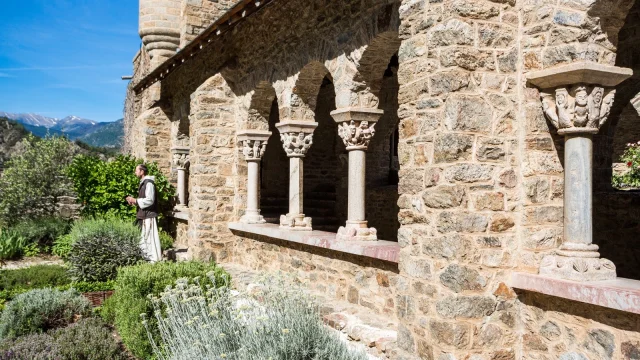 Moine marchant dans le jardin de l’abbaye Saint-Martin du Canigou, devant les arcades en pierre romane sur fond de montagnes.