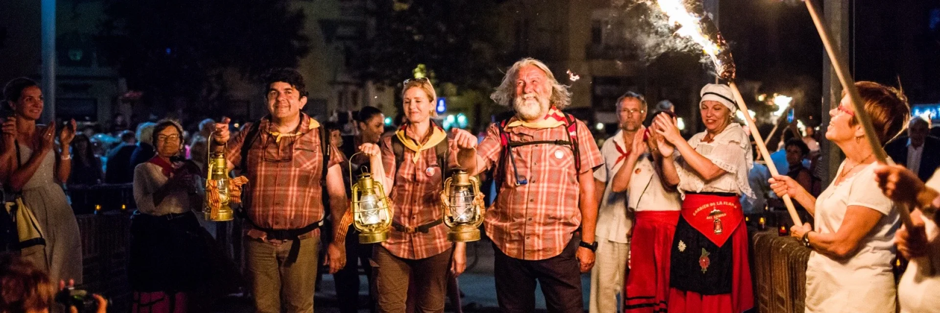Porteurs de la flamme de la Saint-Jean arrivant à Perpignan, tenant des lanternes allumées entourés de spectateurs.