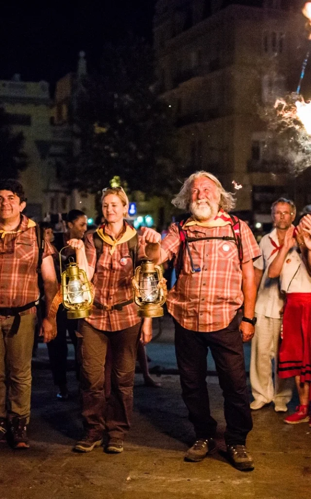 Porteurs de la flamme de la Saint-Jean arrivant à Perpignan, tenant des lanternes allumées entourés de spectateurs.