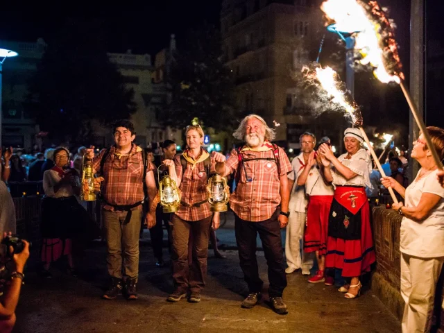 Porteurs de la flamme de la Saint-Jean arrivant à Perpignan, tenant des lanternes allumées entourés de spectateurs.