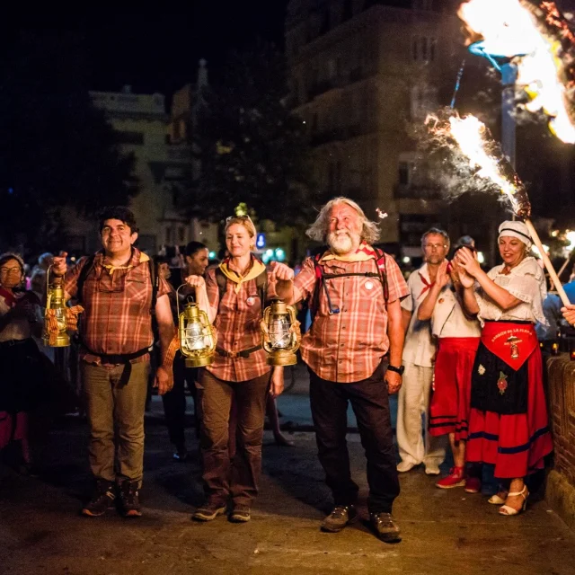 Porteurs de la flamme de la Saint-Jean arrivant à Perpignan, tenant des lanternes allumées entourés de spectateurs.
