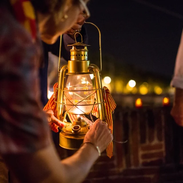 Personnes tenant une lanterne allumée ornée de rubans catalans lors de la fête de la Saint-Jean à Perpignan.