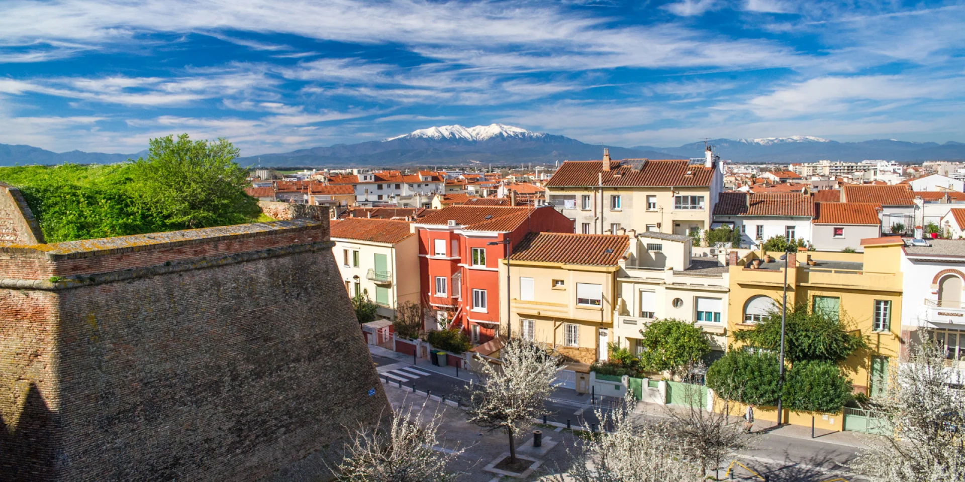 Vue sur les remparts du Palais des Rois de Majorque à Perpignan, avec les toits colorés de la ville et le Canigó enneigé en arrière-plan.