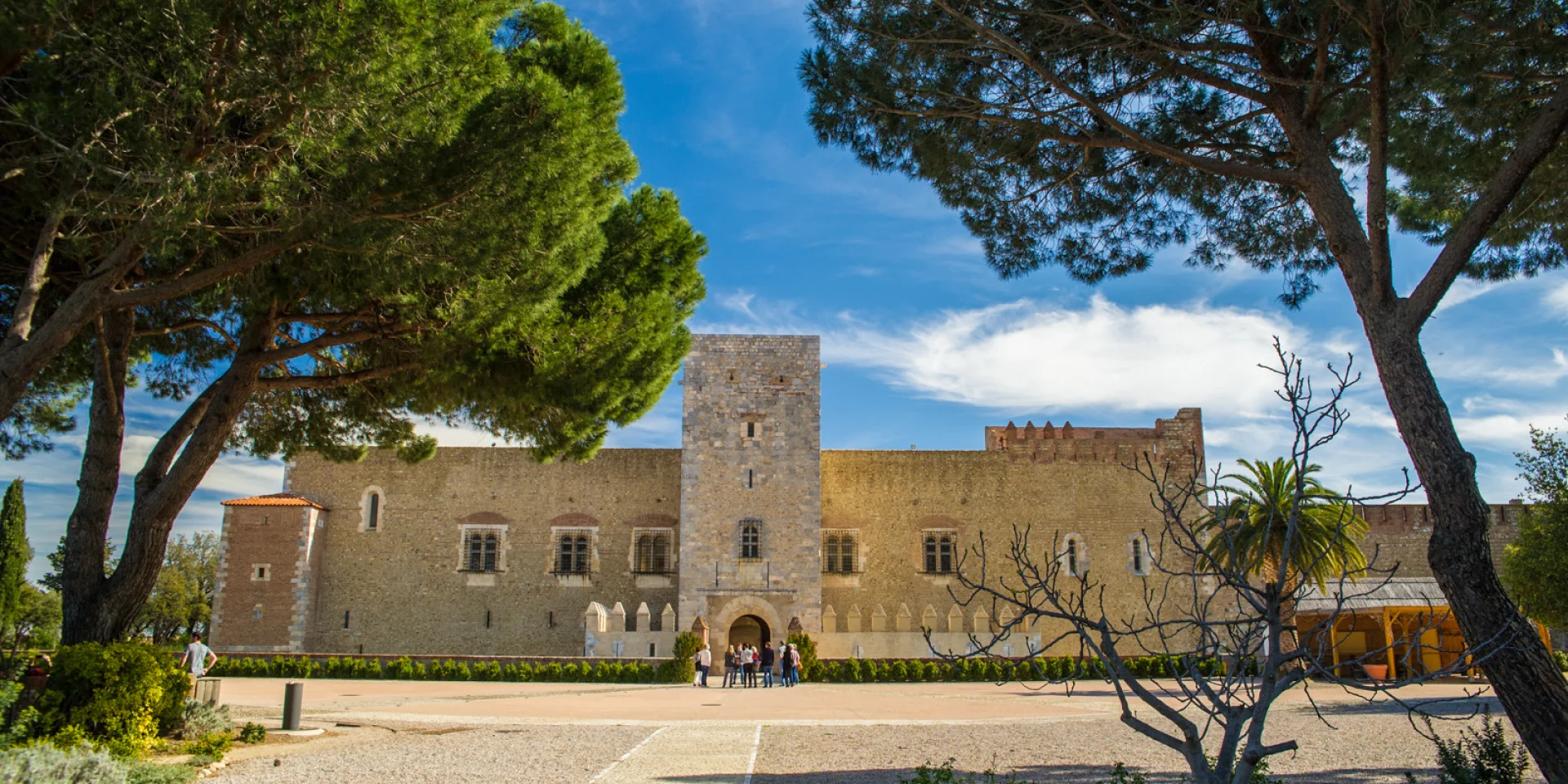 Façade principale du Palais des Rois de Majorque à Perpignan, vue depuis les jardins, encadrée par des pins sous un ciel bleu.