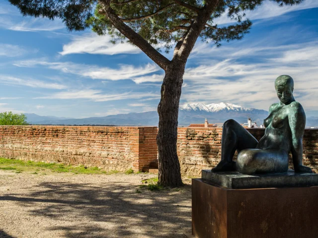 Statue en bronze représentant une femme assise dans les jardins du Palais des Rois de Majorque à Perpignan, avec le Canigó enneigé à l’horizon.