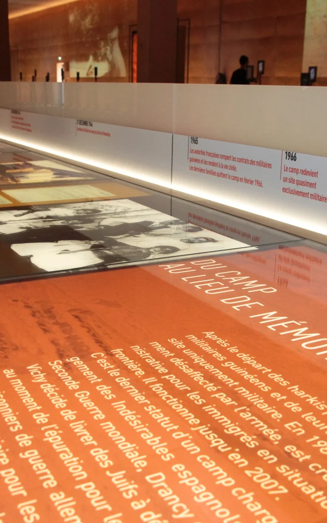 Vitrine d’exposition au Mémorial du Camp de Rivesaltes présentant des textes, des photos d’archives et une frise chronologique retraçant l’histoire du camp.